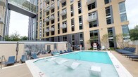 This contemporary multi-story residential building presents a well-designed environment that balances modern aesthetics with functional amenities. The outdoor amenity area serves as a central highlight, featuring a resort-style swimming pool with built-in in-water lounge chairs and a surrounding deck furnished with blue chaise lounges and cushioned seating. The pool area is framed by the building's tall facade, finished in warm beige tones with rows of large windows and private balconies extending from upper floors, creating an enclosed courtyard feel that adds a sense of privacy and scale.

The building's interior common corridors are thoughtfully designed with a consistent, contemporary palette. Wide hallways are lined with smooth, light-gray walls accented by bold teal vertical trim elements that lend a modern edge to the design. Recessed ceiling lighting runs the length of each corridor, ensuring even illumination throughout. The floors are covered in a patterned carpet in shades of gray, teal, and charcoal, reinforcing the building's cohesive color scheme. Unit entry doors are finished in a clean gray tone and feature individual unit numbers accompanied by decorative tile art, providing each residence with a distinct, personalized touch at its entrance.