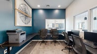 Located at 4037 Lamplighter Dr in North Chesterfield, Virginia, this dedicated business center offers a fully equipped and thoughtfully designed workspace. The room is defined by a bold teal accent wall that adds a vibrant, modern character, contrasting cleanly against the surrounding white walls. Recessed ceiling lighting provides consistent, focused illumination throughout the space.

A continuous dark-surfaced built-in desk wraps along two walls, providing generous workspace for multiple users simultaneously. Several ergonomic mesh-back office chairs are arranged at the workstations, each equipped with desktop computers and monitors. An interior pass-through window connects the room to an adjacent common area, allowing natural light to filter in and maintaining a sense of openness despite the room's compact footprint.

A multifunction printer/scanner is conveniently stationed on a dedicated section of the built-in countertop, making document handling easily accessible. A large framed abstract artwork adorns the teal wall, lending an artistic touch to the otherwise functional environment. The flooring features neutral-toned tile, contributing to the clean, professional aesthetic. A glass-paneled door provides entry and exit while maintaining visual connectivity to the surrounding facility. This space serves as an efficient and well-appointed resource for productivity needs.