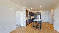 This single-floor unit showcases a contemporary design with a cohesive color palette and thoughtful layout. The kitchen serves as a visual centerpiece, outfitted with slate-blue cabinetry, white quartz countertops, a white subway tile backsplash, and a full suite of stainless steel appliances including a refrigerator, range, microwave, and dishwasher. An island with a bold navy frame and white top provides additional prep and dining space, seamlessly connecting to the open living area. The living space features light hardwood-style flooring that runs throughout the main areas, crisp white walls, and recessed lighting, creating a bright and airy atmosphere. Large windows and a balcony door flood the space with natural light and offer open outdoor views. The bedroom is finished with plush carpet, a ceiling fan, and a generously sized window framing scenic outdoor vistas. The bathroom is outfitted with a navy vanity, white countertop, walk-in shower, and large-format tile flooring, with an adjacent closet providing organized storage. Overall, the unit presents a clean, modern aesthetic with consistent finishes throughout.