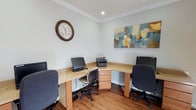 This single-floor business center is designed to provide a functional and comfortable workspace for its users. The room features a built-in wraparound desk with a warm, granite-style surface and natural wood cabinetry, offering multiple dedicated workstations equipped with desktop computers and ergonomic rolling chairs. A multifunction printer is conveniently integrated into the workspace, making it easy to handle a variety of office tasks. The space is finished with crown molding along the ceiling, adding a refined architectural detail, while recessed lighting ensures a bright and focused work environment. Soft, light-toned walls contribute to a calm and professional atmosphere. A decorative abstract canvas painting in muted blues, yellows, and greens adds a touch of color and creativity to the room, while a classic wall clock keeps occupants on schedule. Durable hardwood-style flooring completes the space, offering both practicality and visual warmth. Overall, this business center strikes a balance between efficiency and comfort, making it a well-equipped resource for anyone needing a dedicated space to work, print, or manage professional responsibilities.