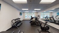 Located at 921 Trailwood Dr in Raleigh, NC, this fitness center offers a well-appointed, single-floor exercise space designed to support a comprehensive workout routine. The room features a drop-panel ceiling with recessed fluorescent lighting that ensures consistent, bright illumination throughout the space. A row of windows along one wall invites natural light and provides views of the surrounding greenery, creating an energizing atmosphere for exercise.

The facility is equipped with a well-rounded selection of cardio machines, including Star Trac treadmills, an elliptical trainer, a stair climber, and a stationary bike, all arranged to allow comfortable movement between stations. A full-length wall mirror runs along one side of the room, visually expanding the space and allowing users to monitor their form during workouts. Free weights are neatly organized on a multi-tier dumbbell rack alongside an adjustable weight bench, catering to strength training needs.

Multiple wall-mounted flat-screen TVs are positioned for easy viewing from various points in the room, adding an entertainment element to workout sessions. The floor is finished with durable rubber flooring in a speckled dark tone, practical for high-activity use. A set of glass-panel doors at the rear of the room provides additional access and further enhances the open, airy feel of this fully equipped fitness facility.