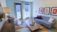 This single-floor space showcases a thoughtfully designed living area that balances modern aesthetics with a warm, inviting atmosphere. The room is anchored by a tufted gray sofa adorned with bold navy and coral accent pillows, complemented by a geometric multi-colored area rug in shades of orange, yellow, and red that adds a dynamic visual element to the polished floor beneath. A low-profile wooden coffee table serves as a functional centerpiece, styled with tasteful decorative objects.

Natural light floods the space through floor-to-ceiling glass doors framed by sheer white curtains, opening directly onto a private balcony furnished with outdoor seating. The balcony access enhances the sense of openness and connectivity to the outside. A sleek brass arc floor lamp provides additional ambient lighting, while framed wall art in red and black tones adds a curated, gallery-like quality to the white walls. The overall palette of soft blues, whites, and warm accent colors creates a cohesive, contemporary feel throughout the space, making it both visually engaging and functionally comfortable.