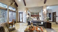 Located in Waxahachie, this single-story space showcases a distinctive rustic aesthetic rooted in Texas Hill Country design. The main living area commands attention with soaring ceilings accented by dark exposed wood beams, creating a dramatic yet welcoming atmosphere. Prominent stacked stone columns frame the open floor plan, adding architectural character and visual depth throughout the space.

The living area is anchored by a tufted leather Chesterfield sofa and a rich hardwood coffee table, complemented by a layered arrangement of southwestern-style rugs that add warmth and color. Travertine tile flooring flows throughout, contributing to the earthy, natural palette of the interior.

A floor lamp and wall sconces provide warm, layered lighting that enhances the cozy ambiance. Natural light filters in through French doors fitted with wooden shutters, offering a connection to the outdoors. Decorative elements including mounted antlers, ceramic vessels, and curated greenery reinforce the space's western-inspired character.

Beyond the living area, the kitchen is visible through the open layout, featuring rich dark wood cabinetry and light countertops. Recessed lighting illuminates the kitchen corridor, ensuring the space remains bright and functional. Overall, the design strikes a refined balance between rustic charm and comfortable, purposeful living.