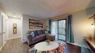 This single-floor residence in Pleasant Hill, California offers a well-organized layout designed for comfortable everyday living. The living room serves as a welcoming central space, featuring dark wood-style flooring, a textured ceiling, and a sliding glass door framed by teal drape panels that opens to a private outdoor balcony with views of surrounding greenery. A wall-mounted HVAC unit provides climate control, and a bold triptych canvas adds a striking focal point above the sectional seating area.

Adjacent to the living room, the dining area benefits from a large vertical-blind window that brings in natural light. A compact white hutch offers display and storage space, complementing the intimate round dining table setup.

The galley-style kitchen is efficiently arranged with white shaker cabinetry, gray countertops, wood-look tile flooring, and stainless steel appliances including a refrigerator, electric range, and dishwasher. Overhead lighting keeps the workspace bright and functional.

The bedroom features plush carpeting, a textured ceiling, and a window dressed with teal curtains, creating a calm, restful environment. The bathroom includes a full tub-shower combination with clean white fixtures and wood-look flooring, maintaining the cohesive style found throughout the unit.