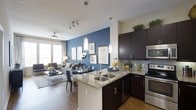 This 1-bedroom, 1-bathroom residence, known as the A3 floor plan, offers a thoughtfully designed open-concept layout that maximizes functionality and style. The kitchen serves as the centerpiece of the living area, featuring rich dark cabinetry, granite countertops, stainless steel appliances, and a peninsula with a double basin sink — ideal for both cooking and casual dining. Pendant lighting above the peninsula adds a refined touch, complementing the track lighting overhead. The kitchen flows seamlessly into the combined dining and living area, which is anchored by a striking blue accent wall adorned with curated artwork. The living space is bathed in natural light from large windows, and a ceiling fan enhances year-round comfort. Warm wood-style flooring runs throughout the main living areas, adding a cohesive and inviting aesthetic. Upper cabinetry in the kitchen extends to the ceiling, providing generous storage, while the open sight lines between the kitchen, dining, and living spaces create an airy, connected atmosphere. The overall design balances contemporary finishes with practical living, making this a comfortable and visually appealing single-floor residence.