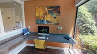 This single-floor business center is thoughtfully designed to provide a functional and comfortable workspace. The main workstation area features an L-shaped granite countertop that wraps along the walls, offering ample desk space for multiple users. Vibrant yellow chairs provide seating at the workstations, which are equipped with an iMac desktop computer, keyboard, and peripheral devices. A bold abstract painting in warm tones of gold, teal, and rust anchors the terracotta-accented accent wall, adding an artistic focal point to the space.

Large windows and a glass sliding door flood the workspace with natural light and offer views of lush, landscaped greenery outside, creating a pleasant and refreshing atmosphere. Interior windows connect the workstation area to an adjacent room, maintaining an open and airy feel throughout.

A secondary room, finished in soft neutral tones with wood-look flooring, houses a full-size multifunction printer and copier alongside a comfortable upholstered accent chair, providing a dedicated area for printing and document handling. The consistent use of wood-look flooring throughout ties the spaces together cohesively. Overall, this business center is a practical and welcoming environment suited for everyday productivity needs.