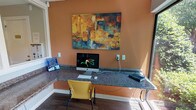 This single-floor business center is thoughtfully designed to provide a functional and comfortable workspace. The main workstation area features an L-shaped granite countertop that wraps along the walls, offering ample desk space for multiple users. Vibrant yellow chairs provide seating at the workstations, which are equipped with an iMac desktop computer, keyboard, and peripheral devices. A bold abstract painting in warm tones of gold, teal, and rust anchors the terracotta-accented accent wall, adding an artistic focal point to the space.

Large windows and a glass sliding door flood the workspace with natural light and offer views of lush, landscaped greenery outside, creating a pleasant and refreshing atmosphere. Interior windows connect the workstation area to an adjacent room, maintaining an open and airy feel throughout.

A secondary room, finished in soft neutral tones with wood-look flooring, houses a full-size multifunction printer and copier alongside a comfortable upholstered accent chair, providing a dedicated area for printing and document handling. The consistent use of wood-look flooring throughout ties the spaces together cohesively. Overall, this business center is a practical and welcoming environment suited for everyday productivity needs.