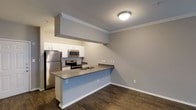 Located in Dallas, TX, this single-floor residence offers a thoughtfully designed open-concept layout that maximizes both functionality and comfort. The kitchen serves as a central highlight, featuring stainless steel appliances including a refrigerator, electric range, and over-the-range microwave. White cabinetry provides ample storage, while a peninsula countertop with a built-in sink creates a natural division between the kitchen and the adjoining living area, also offering casual seating potential. The countertops feature a sleek gray finish that complements the cool-toned cabinetry and the light gray walls throughout. Rich, dark hardwood-style flooring runs continuously across the space, adding warmth and contrast to the lighter walls and cabinetry. Crown molding details the ceiling, lending a refined and polished architectural touch. A soffit above the kitchen cabinetry adds to the defined structure of the space. Flush-mount ceiling lighting ensures consistent illumination throughout. The overall ambiance is clean, modern, and move-in ready, reflecting a practical yet attractive design sensibility well-suited for everyday living in a vibrant urban setting.
