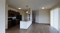 This single-floor residence showcases a thoughtfully designed open-concept layout that seamlessly connects the kitchen and living areas. The kitchen is anchored by a central island topped with granite countertops, providing both a functional workspace and a natural gathering point. Dark, rich cabinetry lines the kitchen walls, offering generous storage, while black appliances — including a full-size refrigerator and range — lend a cohesive, contemporary feel. Pendant lighting hangs above the island, complementing the flush-mount ceiling fixtures that provide warm, even illumination throughout the space. Durable hardwood-style flooring in a warm neutral tone runs continuously across the entire floor plan, tying the spaces together and lending a sense of cohesion and warmth. The open layout transitions smoothly into a spacious living area, offering flexibility in furniture arrangement and use. Neutral-toned walls provide a clean, versatile backdrop that suits a variety of décor styles. A hallway visible from the main living area leads to additional rooms, suggesting a well-organized and practical floor plan. Overall, the space balances everyday functionality with modern design sensibilities.