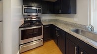 This single-floor residence offers a thoughtfully designed layout with a cohesive blend of modern finishes and functional living spaces. The kitchen is a standout feature, showcasing dark espresso cabinetry with sleek hardware, light granite countertops, and a full suite of stainless steel appliances including a range, over-the-range microwave, refrigerator, and dishwasher. Warm hardwood-style flooring runs throughout the main living areas, adding a polished, inviting feel. The open-concept layout connects the kitchen — complete with a peninsula counter — to the spacious living and dining area, which is brightened by a large window that draws in natural light. A dedicated bedroom offers plush carpeting and additional window views of the surrounding exterior. The bathroom is finished with classic tile work, featuring a patterned mosaic floor, full-height subway-style wall tiles with a decorative accent stripe, and a soaking tub with shower. A practical in-unit laundry closet houses a stacked front-loading washer and dryer, adding convenience to everyday living. The space as a whole maintains a clean, neutral palette throughout, creating a bright and versatile environment ready to accommodate a variety of lifestyles.