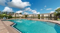 Located at 101 Raceway Pointe Dr in Daytona Beach, Florida, this outdoor amenity area serves as a central gathering space for the residential community. The centerpiece is a generously sized, resort-style swimming pool with crystal-clear turquoise water, accessible via built-in entry steps and framed by a broad, paver-tiled sundeck. The deck is furnished with vibrant yellow Adirondack chairs, providing a relaxed and inviting atmosphere ideal for lounging under the Florida sun. A covered clubhouse structure with large glass doors adjoins the pool area, offering shade and an indoor-outdoor connection for residents. The pool is enclosed by black iron fencing, ensuring safety while maintaining open sightlines to the surrounding landscape. On one side, the pool deck overlooks a tranquil pond with views of the multi-story residential buildings beyond, while mature trees and lush greenery frame the perimeter. A dedicated children's playground with colorful equipment is situated adjacent to the pool area, making this space family-friendly. An outdoor grill station and additional seating areas with umbrella tables complete the amenity offering. The entire space is bathed in abundant natural light beneath Daytona Beach's characteristic bright blue skies, creating a vibrant and welcoming outdoor environment year-round.