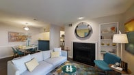 This single-floor residence offers a thoughtfully designed open-concept layout that connects the living area, dining space, and kitchen in a cohesive and functional arrangement. The living room serves as the focal point of the home, anchored by a classic fireplace with a sleek black surround and mantel, above which a large circular mirror adds a striking decorative element. Built-in shelving on the adjacent wall provides convenient display and storage space. The living area is generously proportioned, accommodating a comfortable sofa, glass-top coffee table, and additional seating, all set atop a vibrant geometric-patterned area rug that adds a playful splash of color. The open kitchen is visible from the living space, separated by a teal-accented breakfast bar that doubles as a casual dining counter. The kitchen features standard appliances, including a wall-mounted microwave and range, with light cabinetry providing ample storage. A dedicated dining nook, tucked just off the kitchen, offers space for a full dining set. Neutral wall tones and recessed lighting throughout create a bright, welcoming ambiance.