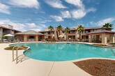 Located at 28851 N Vistancia Blvd in Peoria, Arizona, this outdoor amenity area showcases a generously sized freeform swimming pool as its centerpiece, designed to evoke a resort-style atmosphere under the abundant Arizona sunshine. The pool's organic, curvilinear shape is complemented by a wide, smooth concrete pool deck that provides ample space for lounge chairs, umbrellas, and relaxation areas positioned throughout the perimeter.

A standout feature of the space is the covered cabana pavilion, constructed with stone-clad columns and a sloped tile roof accented by bold red trim. This structure houses a built-in bar with stone tile detailing and high-top seating, offering a shaded gathering spot with direct views of the pool. An outdoor television is mounted within the covered area, adding an entertainment element to the social space.

Tall, mature palm trees are strategically placed around the area, enhancing the desert resort aesthetic while providing natural character and shade. Landscaped planting beds with desert-adapted vegetation line portions of the pool edge, adding color and texture to the hardscape surroundings. The multi-story residential buildings in the warm, earthy stucco tones of Southwestern architecture frame the backdrop, and the space benefits from open sky views and abundant natural light throughout the day.