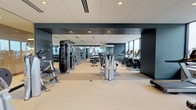 This contemporary fitness center is thoughtfully designed to offer a comprehensive workout experience across a single, well-organized floor. The space is divided into distinct functional zones, each tailored to a specific type of training. A dedicated cardio area lines the floor-to-ceiling windows, where rows of treadmills, ellipticals, and stationary bikes allow users to exercise against a backdrop of expansive city skyline views—an inspiring and energizing feature of the facility. A strength training zone is equipped with a wide selection of resistance machines, cable systems, and a full dumbbell rack, complemented by large wall-mounted mirrors that amplify the sense of space. A glass-walled multipurpose studio with hardwood flooring offers a versatile area for group classes or individual functional training, stocked with medicine balls, resistance bands, and plyo boxes, and fitted with a wall-mounted display screen. The design palette throughout is clean and modern, with muted blue-grey accent walls, neutral carpeted flooring, and a recessed tile ceiling with integrated lighting. The abundance of natural light streaming through the panoramic windows creates an open, airy ambiance that elevates the overall experience of the facility.
