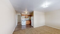This single-floor unit offers a practical and efficient layout, encompassing a living room, kitchen, bedroom, and closet across a modest footprint. The open living and kitchen area seamlessly combines functionality with a neutral, warm aesthetic. The kitchen is equipped with warm-toned wooden cabinetry, dark granite-style countertops, a white electric range, a refrigerator, and a built-in under-cabinet range hood, all set atop durable hardwood-style flooring. The living area transitions naturally from the kitchen and features soft wall-to-wall carpeting, creating a comfortable and inviting atmosphere. The spacious bedroom is carpeted as well, with beige walls and two windows fitted with blinds that allow natural light to filter in; a wall-mounted air conditioning unit provides climate control. A hallway connects the bedroom to a bathroom area featuring a wood-vanity with a single sink and a large mirror, complemented by hardwood-style flooring. Adjacent to the bathroom is a dedicated closet, offering practical storage. Throughout the unit, neutral beige tones and white trim create a clean, cohesive look.