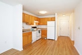 This single-floor residence offers a practical and comfortable layout designed for everyday living. The kitchen is well-equipped with warm honey-toned wood cabinetry extending to the upper walls, providing ample storage space. White appliances including a refrigerator, electric range, dishwasher, and range hood are seamlessly integrated into the layout. Light wood-look flooring runs through the kitchen and connects to an entry hallway, creating a cohesive and easy-to-maintain surface throughout the common areas. The bedrooms feature neutral wall-to-wall carpet and are brightened by windows with views of surrounding greenery, giving each room a calm and airy feel. A textured ceiling adds subtle character to the sleeping quarters. The bathroom is outfitted with a full-size soaking tub and shower combination, accented by a curved curtain rod, alongside standard fixtures and warm wood-look flooring that complements the rest of the home. A dedicated in-unit laundry area includes washer and dryer hookups, wire shelving for added storage, and the same wood-look flooring, making it both functional and organized. The overall layout, as seen in the dollhouse view, is open and efficiently arranged, offering a straightforward flow between all spaces.