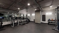 Located at 12012 Wilshire Blvd in Los Angeles, this fitness center offers a well-equipped workout space defined by its industrial aesthetic. The exposed concrete ceiling with visible ductwork, conduit, and piping lends the space a raw, urban character, while recessed and track lighting ensures the area is well-illuminated throughout. Durable rubber flooring covers the entire space, providing both safety and functionality for a wide range of exercises. The cardio zone is outfitted with multiple treadmills, stair climbers, and elliptical machines, all positioned along a full-length mirrored wall that enhances the sense of openness. A dedicated free weights area features a tiered dumbbell rack with a comprehensive weight selection, accompanied by an adjustable bench for strength training. Kettlebells of varying weights are neatly organized on a separate rack, complemented by a cable and functional training machine for resistance workouts. A wall-mounted flat-screen television adds convenience for entertainment during workouts. Exposed concrete columns punctuate the floor plan, reinforcing the industrial design language. The space is accessible via glass-paneled doors and is situated adjacent to a mail room, suggesting its role as a shared amenity within a larger building.