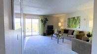 This single-floor living space offers a well-appointed, open layout centered around a generously sized living area. The room features plush wall-to-wall carpeting, contributing to a comfortable and inviting atmosphere. A key highlight is the large sliding glass door that floods the space with abundant natural light while providing direct access to a private outdoor patio, where a small seating area is visible beyond the yellow exterior wall.

The living area is thoughtfully arranged with a neutral-toned sofa accented by green and purple decorative pillows, complemented by a sleek glass-top coffee table. A flat-screen television is positioned on a low media console in the corner, flanked by a tall indoor fiddle-leaf fig plant that adds a natural, organic element to the space. Green patterned curtains frame the sliding door, tying together the room's nature-inspired color palette.

A wall-mounted sconce and a floor lamp provide warm ambient lighting, while botanical artwork adorns the wall, enhancing the cohesive aesthetic. The textured ceiling adds a classic architectural detail throughout the space.