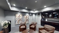 This well-appointed single-floor space offers a refined and contemporary atmosphere designed for comfort and leisure. The lounge area serves as the focal point, showcasing rich brown leather armchairs arranged around a low central table, creating an inviting setting for conversation or relaxation. The walls are finished in a cool, sophisticated grey tone that lends the space a modern, polished character. Overhead, a series of recessed lights are set into a dark ceiling, providing warm, even illumination throughout the room. Two large-scale abstract artworks hang prominently on the wall, featuring bold circular brushstrokes in muted tones with accents of red and gold, adding a gallery-like quality to the interior. A built-in dark wood cabinetry unit occupies one corner of the room, offering both storage and functionality, with a microwave and display shelving integrated into the design. A wall-mounted speaker and ceiling-inset audio components suggest the space is equipped for an enhanced entertainment experience. The overall ambiance is sleek and intimate, balancing modern design sensibilities with warm, comfortable furnishings to create a space that is both visually engaging and highly functional.