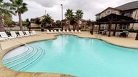 This outdoor pool area is a thoughtfully designed recreational space that serves as a central amenity for the surrounding residential community. The centerpiece is a generously sized swimming pool with a freeform shape and clear, blue-tinted water, complemented by an adjacent spa in certain views — offering options for both active swimming and relaxation. The pool deck is finished in light-toned concrete and lined with multiple lounge chairs, providing ample space for sunbathing and outdoor leisure.

A covered entry pavilion anchors one side of the pool area, featuring dark wood-stained structural columns, stucco exterior walls, and warm wood-framed doors with glass panels. Wall-mounted light fixtures and potted planters add character to this transitional space, which also displays pool rules signage for resident reference. Large sliding glass doors in the pavilion provide a visual connection between the interior and the pool deck.

The pool area is enclosed by fencing and framed by mature trees and palm trees, lending a resort-like atmosphere. A shaded pergola with seating on one end offers a comfortable retreat from the sun. The surrounding multi-story residential buildings are visible from the pool deck, reinforcing the community-oriented nature of this amenity. The space is designed to be both functional and inviting for everyday outdoor enjoyment.