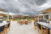 Located at 1 Ridge Rd in Auburn, Massachusetts, this generously sized outdoor amenity space offers a well-appointed and thoughtfully designed environment for relaxation and recreation. The space is anchored by a sweeping stone-paved patio that provides ample room for multiple seating arrangements, including a central lounge area furnished with teak armchairs and cushioned seats ideal for gathering. Pergola structures frame the patio edges, adding architectural definition and shade. Adjacent to the patio, a vibrant green lawn area serves as an open recreational zone, complete with cornhole boards and room for various outdoor activities. A dedicated fire pit lounge area features a circular concrete fire pit surrounded by Adirondack chairs, creating an inviting setting for evening gatherings. Outdoor grilling stations are conveniently positioned nearby. The perimeter of the space is thoughtfully landscaped with ornamental grasses, evergreen shrubs, and deciduous trees that provide seasonal color and visual interest, all enclosed by a sleek black iron fence. The exterior building facades feature neutral lap siding with gable rooflines and large windows. The surrounding multi-story residential buildings are visible in the backdrop, reinforcing the communal character of this well-equipped outdoor retreat.
