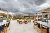 Located at 1 Ridge Rd in Auburn, Massachusetts, this generously sized outdoor amenity space offers a well-appointed and thoughtfully designed environment for relaxation and recreation. The space is anchored by a sweeping stone-paved patio that provides ample room for multiple seating arrangements, including a central lounge area furnished with teak armchairs and cushioned seats ideal for gathering. Pergola structures frame the patio edges, adding architectural definition and shade. Adjacent to the patio, a vibrant green lawn area serves as an open recreational zone, complete with cornhole boards and room for various outdoor activities. A dedicated fire pit lounge area features a circular concrete fire pit surrounded by Adirondack chairs, creating an inviting setting for evening gatherings. Outdoor grilling stations are conveniently positioned nearby. The perimeter of the space is thoughtfully landscaped with ornamental grasses, evergreen shrubs, and deciduous trees that provide seasonal color and visual interest, all enclosed by a sleek black iron fence. The exterior building facades feature neutral lap siding with gable rooflines and large windows. The surrounding multi-story residential buildings are visible in the backdrop, reinforcing the communal character of this well-equipped outdoor retreat.