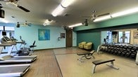 This single-floor fitness room offers a practical and comfortable space for exercise. The room features a combination of smooth flooring and carpet tile sections, delineating different workout zones. The walls are painted in a soft aqua-green tone, accented by a deep teal feature wall that adds visual interest and depth. A vibrant abstract canvas painting adorns one wall, contributing an artistic touch to the functional space.

Overhead, two ceiling fans provide consistent airflow throughout the room, complementing the fluorescent lighting that ensures bright, even illumination. The fitness area is equipped with a range of exercise equipment, including a treadmill, weight bench, and a dumbbell rack positioned near a large wall mirror, which enhances the sense of space and allows users to monitor their form during workouts.

A small seating area with two chartreuse upholstered chairs offers a rest space near the entrance. A standard door provides access to the room, alongside posted informational signage. The overall ambiance is clean and functional, designed to support a variety of workout routines in a well-maintained, community-style fitness environment.