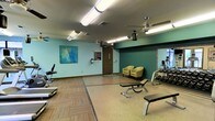 This single-floor fitness room offers a practical and comfortable space for exercise. The room features a combination of smooth flooring and carpet tile sections, delineating different workout zones. The walls are painted in a soft aqua-green tone, accented by a deep teal feature wall that adds visual interest and depth. A vibrant abstract canvas painting adorns one wall, contributing an artistic touch to the functional space.

Overhead, two ceiling fans provide consistent airflow throughout the room, complementing the fluorescent lighting that ensures bright, even illumination. The fitness area is equipped with a range of exercise equipment, including a treadmill, weight bench, and a dumbbell rack positioned near a large wall mirror, which enhances the sense of space and allows users to monitor their form during workouts.

A small seating area with two chartreuse upholstered chairs offers a rest space near the entrance. A standard door provides access to the room, alongside posted informational signage. The overall ambiance is clean and functional, designed to support a variety of workout routines in a well-maintained, community-style fitness environment.