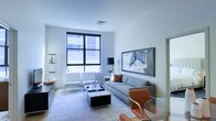 This single-floor space showcases a refined, contemporary design with a bright and airy atmosphere. The living area is anchored by large, black-framed floor-to-ceiling windows that flood the room with natural light, offering views of the surrounding urban streetscape. The interior palette is kept crisp and neutral, with white walls and light flooring creating a seamless, open feel throughout.

The living area is thoughtfully furnished with a patterned gray sofa accented by warm rust-toned throw pillows, complemented by sleek chrome-legged seating and a mirrored coffee table that enhances the sense of space. A large framed art piece adorns the wall, adding a sophisticated focal point to the room. A tan leather accent chair and glass-topped surface introduce warmth and variety to the otherwise cool, modern palette.

Recessed ceiling vents and clean architectural lines speak to the careful attention to detail in the space's design. Built-in shelving with glass accents is visible to one side, offering both functional storage and a display element. The overall ambiance is polished and professional, blending modern minimalism with carefully chosen furnishings to create a space that feels both stylish and welcoming.