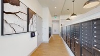 Located at 500 River Knoll Way in Columbus, Georgia, this dedicated mail and package room offers a clean, modern environment designed for efficient resident mail management. The single-floor space features light wood-look flooring throughout, crisp white walls, and notably high ceilings that lend an open, airy feel well beyond its compact footprint. Natural light filters in through transom-style windows dressed with sheer white curtains, complementing the recessed ceiling lights and stylish industrial-style pendant fixtures that illuminate the space warmly. Lining the walls are extensive banks of dark bronze individual mailboxes, numbered and organized for easy access. Adjacent to these are modern smart parcel lockers in a matching dark finish, providing secure package retrieval for residents. A digital access panel is conveniently mounted near the entry, enabling keyless and app-based entry. The walls are further enhanced by large-scale framed abstract artwork in neutral tones, adding an elevated aesthetic touch to an otherwise functional space. The overall ambiance is polished and welcoming, combining practical utility with thoughtful design details that reflect the community's commitment to resident convenience and contemporary living standards.