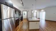 This well-appointed single-floor residence offers a thoughtfully designed open-concept layout that seamlessly connects the kitchen and living areas. The kitchen is a standout feature, showcasing dark espresso cabinetry, granite countertops, a mosaic tile backsplash, and a full suite of stainless steel appliances including a refrigerator, range, and over-the-range microwave. A large central island with a built-in sink and pendant lighting above provides additional prep space and a natural gathering point. Track lighting runs throughout the kitchen and living area, adding both function and a modern aesthetic. Rich hardwood-style flooring extends across the main living spaces, complementing the clean, neutral color palette. The spacious living area is filled with natural light from a sliding glass door with vertical blinds and an adjacent window offering outdoor views. The bedroom features plush carpeting, a ceiling fan, and its own window for natural light. The bathroom is finished with matching granite countertops, dark wood cabinetry, a large framed mirror, and hardwood-style flooring. A dedicated laundry area with a full-size washer and overhead shelving adds everyday convenience, completing this modern and functional residence.