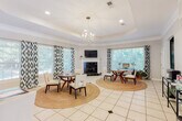 Located at 151 Grande View Dr in Biloxi, Mississippi, this single-floor space offers a refined and functional layout designed to impress. The welcoming entryway greets visitors with soaring ceilings, a striking orb chandelier, large arched double doors, and a soft coastal-toned runner over light tile flooring — setting an inviting tone from the moment you step inside.

The main gathering area is expansive and airy, anchored by a tray ceiling with crown molding and the same stylish orb chandelier. Multiple seating arrangements, each centered on round jute rugs, provide a warm and organized layout ideal for meetings or social gatherings. A white-mantled corner fireplace with black tile surround serves as a focal point, complemented by a wall-mounted television above. Numerous large windows dressed in bold teal ikat-patterned curtains flood the space with natural light and views of lush surrounding greenery.

A private office area accessible via a glass-paned French door offers a more intimate setting, complete with a tray ceiling, crown molding, a dedicated workstation, and additional natural light from a side window. The consistent light tile flooring, neutral gray walls, and cohesive decor throughout create a polished, professional yet comfortable ambiance.