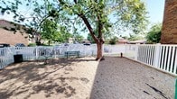 This inviting outdoor space is designed as a dedicated pet or dog park area, thoughtfully enclosed by a classic white picket fence that defines its boundaries while maintaining an open, welcoming feel. At the heart of the space stands a large, mature tree whose expansive canopy provides generous natural shade across the gravel-covered ground below, creating a comfortable environment even on sunny days. The dappled light filtering through the tree's leafy branches lends the area a pleasant, relaxed ambiance. A green wooden bench is positioned nearby, offering a comfortable spot for visitors to sit and relax while keeping watch. Decorative metal accents, including what appears to be a bicycle wheel ornament, add a touch of character and charm to the space. The ground is finished with a gravel and decomposed granite surface, which is both practical and low-maintenance. Views beyond the fence reveal a quiet residential neighborhood with mature trees and nearby buildings visible in the background, contributing to a sense of community. The overall atmosphere is casual, functional, and well-suited for outdoor leisure and pet activity.