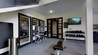 This fitness center offers a thoughtfully designed single-floor layout built to support a comprehensive workout experience. The space features a neutral color palette with crisp white walls contrasted by bold black-framed glass doors and partitions, lending it a sleek, modern aesthetic. The main workout area is furnished with professional-grade strength training machines, including a leg press/curl station and an adjustable weight bench, providing ample options for resistance training. A well-stocked dumbbell rack lines one wall, offering a wide range of free weights neatly organized for easy access. A wall-mounted flat-screen television is positioned prominently, allowing users to stay entertained or informed during their workouts. The black-framed glass partitions create a visual connection to an adjacent exercise room, which is visible through the panels and appears to contain additional equipment, including cardio machines. Recessed and overhead lighting throughout ensures the space is well-lit, while the polished concrete-style flooring adds a durable, gym-appropriate finish. The layout is open and uncluttered, allowing for easy movement between stations. Overall, the space delivers a clean, functional, and professional environment suited for a variety of fitness routines.