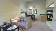 This single-floor unit is designed with an open-concept layout that seamlessly connects the living, dining, and kitchen areas. The living room features plush carpeting, accented by a colorful striped area rug, and is furnished with a comfortable sofa and coffee table. A front-facing window allows natural light to fill the space, creating a bright and welcoming atmosphere. Wall art and decorative shelving add personal touches throughout.

A notable architectural feature is the vaulted ceiling in the living and dining area, which enhances the sense of openness and volume. A ceiling fan with a light fixture is centrally mounted, providing both airflow and overhead illumination. The dining area accommodates a small table and chairs, flanked by two framed mirrors that reflect light and add visual depth to the space.

Adjacent to the dining area, the kitchen features dark countertops and wood-toned cabinetry, offering practical storage and prep space. The entry foyer provides a defined transition into the main living area, complete with a welcome mat. Overall, the unit presents a cozy yet spacious feel with a neutral color palette throughout.