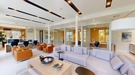 Located at 3300 Boutwell Rd in Lake Worth Beach, Florida, this thoughtfully designed single-floor amenity space offers a seamless blend of lounge and co-working functionality. The open-plan layout is anchored by a spacious living area furnished with a large, light-gray sectional sofa, a natural wood coffee table, and vibrant cognac leather accent chairs — creating a welcoming, residential-style atmosphere with a modern edge.

A bold cobalt blue accent wall adds a striking focal point to the lounge area, complemented by curated artwork and contemporary arc floor lamps. The ceiling features a distinctive design with dark linear recessed lighting strips set against a crisp white backdrop, adding architectural dimension throughout.

Along the perimeter, a row of glass-enclosed private workstations lined with warm amber-toned wall panels provides semi-private work areas equipped with built-in desks and ergonomic seating. An open co-working station with a long shared desk and task chairs offers additional workspace options.

Large windows and glass double doors along the exterior wall flood the space with natural light and offer views of the surrounding greenery. Light-toned tile flooring runs throughout, tying together the clean, contemporary aesthetic. The overall ambiance is professional yet inviting, purpose-built to support both productivity and relaxation.