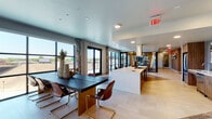 This contemporary single-floor amenity space is thoughtfully designed to blend functionality with modern aesthetics. The open-plan layout connects multiple distinct zones, creating a seamless flow throughout the space. Large floor-to-ceiling windows and glass-panel doors flood the interior with natural light while framing dynamic urban views of the surrounding cityscape, including elevated roadways and the bustling streetscape below.

The fully equipped kitchen is a standout feature, outfitted with stainless steel appliances including a full-size refrigerator, built-in ovens, an induction cooktop, and a sleek island with a pendant hood — all finished with warm wood cabinetry and light stone countertops. Adjacent to the kitchen, the open dining area accommodates a bold dark wood communal table paired with leather and brass cantilever chairs.

The lounge area offers generous seating with deep leather sofas arranged around a wall-mounted TV and a contemporary linear fireplace, all beneath a striking coffered ceiling fitted with geometric hexagonal pendant lighting. A wood slat partition elegantly delineates this space. A secondary lounge corridor features warm wood-paneled walls, plush blue accent chairs, a patterned area rug, and access to private meeting or office rooms. French doors lead to an outdoor balcony, extending the space's connection to the outdoors.