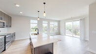 This single-floor residence showcases a clean, contemporary design characterized by a neutral color palette, light-toned hardwood-style flooring, and an abundance of natural light. The open-concept living and kitchen area serves as the centerpiece of the unit, where a large island with a white quartz countertop, undermount sink, and matte black pendant lighting anchors the space. The kitchen is fully equipped with stainless steel appliances, including a French door refrigerator, electric range, and over-the-range microwave, complemented by rich, dark wood cabinetry extending to the ceiling for ample storage.

The living area flows seamlessly toward a wall of windows and a sliding glass door that opens to a private balcony, offering outdoor views and filling the space with natural light. Bedrooms feature plush carpeting, ceiling fans, and generously sized windows that frame views of the surrounding neighborhood. The bathroom is finished with a dark-framed vanity mirror, quartz countertop, undermount sink, wood-toned cabinetry, and a glass-enclosed walk-in shower. A hallway connects the rooms, lined with contemporary wood-panel interior doors, contributing to the unit's cohesive modern aesthetic throughout.