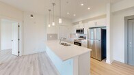 Located in the Nolan Hill community of Calgary, AB, this 2-bedroom, 2-bathroom unit offers a well-designed single-floor layout that makes efficient use of its space. The open-concept kitchen and living area serves as the heart of the home, featuring a large central island with a quartz countertop and undermount sink, white shaker-style cabinetry, a classic tile backsplash, and a full suite of stainless steel appliances including a refrigerator, electric range, and microwave. Pendant lighting above the island adds a modern touch, while recessed pot lights illuminate the space throughout. Light-toned luxury vinyl plank flooring runs continuously across all areas, lending a clean and cohesive aesthetic. The living room opens to a private balcony through a glass-panel door, with large windows framing views of the surrounding green hillside. Both bedrooms are bright and well-proportioned, also offering scenic views of the open landscape. The primary bedroom is served by a full ensuite bathroom with a glass-enclosed walk-in shower, while the second bathroom features a soaker tub and shower combination with a sleek vanity and mirror. A dedicated closet and hallway complete the functional layout of this modern, light-filled home.