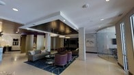 This single-floor lobby space showcases a refined, contemporary design that immediately conveys a sense of upscale hospitality. The most striking architectural feature is the bold coffered ceiling detail, finished in deep espresso tones and framed by crisp white surrounds, which defines the central seating zone and draws the eye downward to the carefully curated furnishings below. A trio of drum-style pendant lights hangs within this recessed ceiling feature, casting warm, layered illumination throughout the space.

The lounge area is anchored by a rich navy and gold area rug, atop which plush purple velvet accent chairs and neutral-toned sofas are arranged around glass-top coffee tables, offering both visual contrast and inviting comfort. A built-in media console with cabinetry provides a functional focal point along one wall. Structural columns clad in light stone tile add architectural interest while defining the spatial layout. To one side, a curved glass enclosure—likely an elevator or vestibule—introduces a sleek, modern element. Artwork and subtle wall accents complete the polished aesthetic, contributing to an overall ambiance that is both professional and welcoming.