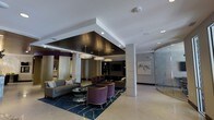 This single-floor lobby space showcases a refined, contemporary design that immediately conveys a sense of upscale hospitality. The most striking architectural feature is the bold coffered ceiling detail, finished in deep espresso tones and framed by crisp white surrounds, which defines the central seating zone and draws the eye downward to the carefully curated furnishings below. A trio of drum-style pendant lights hangs within this recessed ceiling feature, casting warm, layered illumination throughout the space.

The lounge area is anchored by a rich navy and gold area rug, atop which plush purple velvet accent chairs and neutral-toned sofas are arranged around glass-top coffee tables, offering both visual contrast and inviting comfort. A built-in media console with cabinetry provides a functional focal point along one wall. Structural columns clad in light stone tile add architectural interest while defining the spatial layout. To one side, a curved glass enclosure—likely an elevator or vestibule—introduces a sleek, modern element. Artwork and subtle wall accents complete the polished aesthetic, contributing to an overall ambiance that is both professional and welcoming.