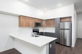 This single-floor residence features a thoughtfully designed layout connecting the main living areas with ease. The kitchen is a standout space, equipped with a full suite of stainless steel appliances, including a refrigerator, electric range, built-in microwave, and dishwasher. Cabinetry combines wood-toned upper cabinets with sleek grey lower units, complemented by white quartz countertops and a glass tile backsplash. A peninsula with an integrated sink and additional seating potential opens toward the living area, which is accented by soft teal wall tones and durable hardwood-style flooring. Recessed lighting illuminates the kitchen and living space, while a window brings in natural light from outside. The bedroom features neutral carpet flooring, a large window overlooking a balcony railing and greenery, and a baseboard heater. A utility panel is conveniently accessible in the hallway connecting the bedroom to the kitchen. The bathroom is finished with a full tub-shower combination with tile surround, wood-toned vanity cabinetry, white countertop, and hardwood-style flooring, presenting a clean and functional design throughout.