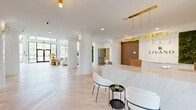 The Livano clubhouse is a thoughtfully designed, single-floor amenity space that seamlessly blends modern elegance with warm, organic elements. Upon entry, guests are greeted by a stunning reception area anchored by a marble-clad desk beneath brass pendant lighting, flanked by white-tiled cylindrical columns and light hardwood floors laid in a herringbone pattern. A living moss wall and warm wood-paneled accents add a natural counterpoint to the crisp white palette throughout.

The expansive lounge area features soaring double-height ceilings and floor-to-ceiling glass walls that flood the interior with natural light and offer views of the lush outdoor surroundings, including a pool and palm trees. Comfortable seating groupings, a wood-paneled media wall, and sculptural rattan lighting create an inviting, resort-like atmosphere.

A dedicated dining zone showcases an ornate coffered ceiling with integrated acoustic panels and a striking chandelier, centered around an oval wood dining table with upholstered seating. Nearby, a fully outfitted kitchen finished in deep green cabinetry provides a bold contrast to the otherwise neutral interior. Arched doorways with dark wood surrounds and white tile columns guide the flow between zones, lending architectural character throughout this refined and functional communal space.