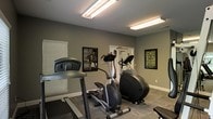 This single-floor fitness space is designed to provide a focused and functional workout environment. The room features neutral gray walls paired with white trim and baseboards, creating a clean, modern aesthetic. Bright fluorescent overhead lighting ensures the space is well-illuminated throughout, supporting visibility during any hour of the day.

The fitness area is outfitted with essential cardio equipment, including a treadmill, an elliptical cross-trainer, and a stationary cycle, arranged to maximize floor space and ease of movement between machines. Durable, low-pile carpet covers the floor, offering both comfort and stability during exercise sessions.

Framed motivational artwork adorns the walls, adding an energizing and purposeful atmosphere to the room. A set of glass-panel doors leads to an adjacent area, allowing for natural light to filter in and providing a sense of openness. Windows fitted with blinds offer additional light control and privacy as needed.

The overall ambiance of the space is practical and inviting, thoughtfully arranged to encourage regular physical activity. The combination of quality equipment, motivational décor, and efficient layout makes this fitness room a well-rounded facility suited for a variety of cardio-focused workout routines.