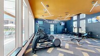 This fitness center in Baton Rouge, Louisiana, is a thoughtfully designed, single-floor facility divided into two distinct workout areas. The main exercise room makes an immediate visual impact with its striking bold blue walls, a vivid amber-orange ceiling, and geometric linear LED lighting that crisscrosses overhead. A motivational "Train Hard" mural is prominently displayed on the feature wall, reinforcing the space's energetic atmosphere. This primary area houses a comprehensive selection of equipment, including elliptical trainers, treadmills, a stair climber, weight benches, a cable machine, a power rack, and a full dumbbell rack. Wall-mounted flat-screen TVs are positioned throughout for entertainment during workouts. Large floor-to-ceiling windows and oversized glazed panels flood the space with natural light while offering views of the surrounding outdoor amenity areas. The flooring throughout features a stylish patterned carpet tile in blue, grey, and tan tones. A secondary room, distinguished by its bright teal ceiling and soft neutral walls, serves as a dedicated studio-style space ideal for cycling and rowing, and is similarly bathed in natural light through tall vertical windows. Together, both rooms deliver a vibrant, functional, and well-lit environment suited for a full range of fitness activities.