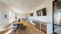 The Parklands Clubhouse, located on the third floor in Ventura, California, is a thoughtfully designed community gathering space that blends modern aesthetics with functional versatility. The single-floor layout flows seamlessly across distinct zones, all unified by warm wood-look flooring and a bright, neutral color palette.

The central hub of the space features a generous communal dining area anchored by a solid wood table with bench seating, situated alongside a well-appointed kitchenette. The kitchenette includes stainless steel appliances—featuring a built-in microwave, refrigerator, and sink with modern fixtures—white cabinetry, and a large-format white tile backsplash. Multiple wall-mounted flat-screen televisions serve both the kitchen and lounge areas.

A comfortable lounge area is defined by a sectional sofa, rustic wood coffee tables, and a textured dark accent wall, while additional seating nooks feature teal accent chairs and round tables bathed in natural light from large windows. Recessed lighting and pendant fixtures throughout provide a well-lit atmosphere.

French doors open to a balcony offering elevated views of the surrounding Ventura neighborhood, and high-top bistro seating near the doors creates an ideal casual gathering spot. The overall ambiance is contemporary, welcoming, and well-suited for community events and social interaction.