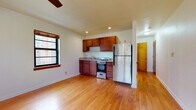 This single-floor unit offers a practical and inviting layout centered around an open-plan living and kitchen area. Rich hardwood flooring flows throughout the main space, lending warmth and continuity to the interior. The kitchen is efficiently designed with warm-toned wood cabinetry, granite-style countertops, a stainless steel refrigerator, and a gas range with oven — providing a fully functional cooking space without compromising the openness of the living area. Recessed lighting and a ceiling fan contribute to a well-lit, comfortable atmosphere.

The living area is generously proportioned, with multiple windows framed in dark wood trim that allow natural light to fill the room. A classic radiator beneath one window hints at the building's established character. The open layout between the kitchen and living area creates a flexible, airy feel ideal for both everyday living and entertaining.

The bathroom features clean white subway tile on the tub surround, accented by a decorative mosaic border in earth tones of brown, rust, and gray — adding a stylish touch to an otherwise crisp, classic design. A hallway connects the main living space to additional areas, including a secondary room accessible through a warm wood-toned door, rounding out this well-appointed, single-level space.