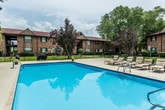 This outdoor pool area offers a welcoming communal retreat surrounded by lush, mature trees and a classic brick building backdrop. The centerpiece is a generously sized swimming pool with clear, blue water, accessible via built-in entry rails and steps. The expansive concrete deck surrounding the pool provides ample space for relaxation and socializing, furnished with multiple lounge chairs for sunbathing as well as a dining-height table and chairs shaded by a bright teal umbrella — ideal for outdoor gatherings. A wooden privacy fence lines one side of the deck, defining the space and adding a sense of enclosure. Conveniently located on-site are separate men's and women's restroom facilities, accessible directly from the pool deck. An additional utility room provides storage space for equipment and maintenance supplies. The open-air setting benefits from natural light throughout the day, with the surrounding greenery offering partial shade and a pleasant, park-like ambiance. Overall, the pool area is a functional and inviting outdoor amenity designed to serve as a central social and recreational hub.