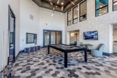 Located at 13220 Foster St in Overland Park, Kansas, this contemporary community clubhouse offers a thoughtfully designed, multi-zone social space ideal for recreation and relaxation. The space is anchored by a dramatic game room boasting soaring double-height ceilings, striking geometric hexagonal tile flooring in varying tones, and a sleek pool table beneath an eye-catching angular pendant light fixture. Clerestory windows flood the area with natural light, while a wall-mounted flat-screen TV and cozy seating nook complete the entertainment-ready atmosphere. Adjacent to the game room, a spacious lounge area features plush, channel-tufted sofas in soft blue-gray tones, layered with geometric-patterned accent walls and warm wood flooring, creating an inviting retreat for socializing. The fully equipped kitchen and dining area showcases white subway tile backsplash, flat-front white cabinetry, a generous island with bar seating beneath bold black conical pendant lights, and a round dining table surrounded by comfortable chairs — all bathed in natural light from oversized windows overlooking an outdoor courtyard. Throughout the space, a cohesive modern design palette of neutral tones, teal accents, and contemporary furnishings ties each zone together seamlessly, offering residents a versatile and stylish communal environment.