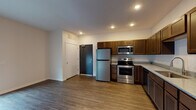 This single-floor unit presents a clean, functional layout designed for everyday living. The open-concept kitchen and living area form the heart of the space, finished with warm wood-style luxury vinyl plank flooring that extends throughout the main areas. The kitchen is well-appointed with rich, dark-stained shaker-style cabinetry offering generous upper and lower storage, complemented by light granite-style countertops. A full suite of stainless steel appliances is included, featuring a refrigerator with top freezer, an electric range, over-the-range microwave, and a dishwasher. A double-basin sink with a modern pull-down faucet anchors the countertop workspace. Recessed lighting illuminates both the kitchen and living areas efficiently. The living space is open and versatile, with neutral white walls providing a clean backdrop. A dedicated bedroom area includes a walk-in closet fitted with wrap-around hanging rods and shelving, carpeted for comfort underfoot. The bathroom features a full soaking tub with a tiled surround and built-in corner shelving. A bold dark entry door adds a contemporary touch to the unit's overall aesthetic.